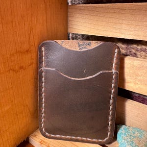 May include: A dark brown leather card holder with two pockets and white stitching. The edges are rounded, and the leather has a slightly textured appearance. The card holder is displayed on a wooden surface.