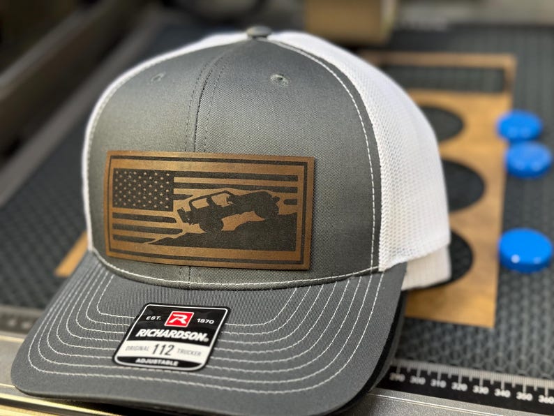 May include: A gray and white trucker hat with a brown leather patch featuring an American flag and a silhouette of a Jeep driving over a hill. The patch has the text "EST. 1970 RICHARDSON 112 ADJUSTABLE" on it.