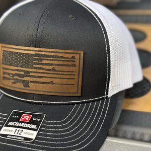 May include: A black and white trucker hat with a brown leather patch featuring a silhouette of an American flag with rifles. The patch has the text "EST. 1970 RICHARDSON. ORIGINAL TRUCKER 112 ADJUSTABLE" on it.