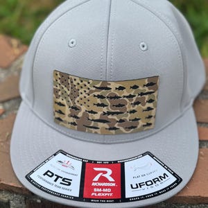 May include: A light gray baseball cap with a flat brim. The cap features a rectangular patch with a camouflage design of fish and stars, resembling a flag. The brim has a sticker with the text "RICHARDSON SM-MD FLEXFIT".