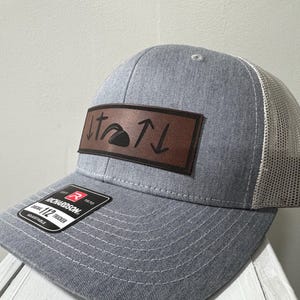 May include: A gray and white trucker hat with a brown leather patch that says "It's a ↑↓" with a mountain icon in the middle.