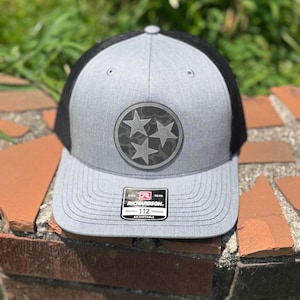 May include: Gray and black trucker hat with a Tennessee state flag emblem. The hat features a gray front panel, a black mesh back, and a curved brim. The emblem is a circular patch with three stars. The hat is adjustable.