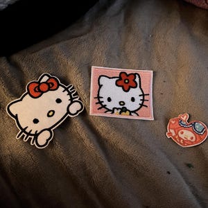 May include: Three embroidered patches featuring Hello Kitty and related characters. One patch is a white Hello Kitty with a red bow, another is a square patch with Hello Kitty in a pink frame, and the third is a heart-shaped patch.