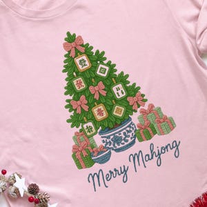 May include: Pink t-shirt featuring a Christmas tree design made of green branches, pink bows, and Mahjong tiles. The tree sits in a blue and white pot, surrounded by wrapped gifts. The text "Merry Mahjong" is written in cursive.