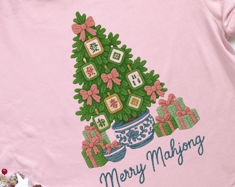 May include: Pink t-shirt with a Christmas tree design made of green branches, pink bows, and Mahjong tiles. The tree is in a blue and white pot, surrounded by wrapped gifts. The text "Merry Mahjong" is written in cursive.