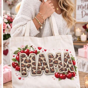 Op de afbeelding: Crèmekleurige draagtas met het woord "MAMA" in letters met luipaardprint. Het ontwerp bevat rode kersen, groene bladeren en kleine rode hartjes. De tas wordt vastgehouden door een persoon die een witte trui draagt.