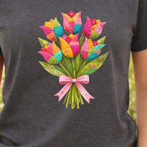 Op de afbeelding: Donkergrijs T-shirt met een kleurrijk boeket tulpen. De tulpen zijn in patchwork-stijl met roze, gele, blauwe en groene tinten. Een roze en wit geruit lint bindt het boeket samen. Het T-shirt heeft een ronde hals.