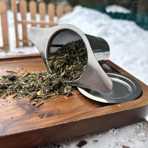 May include: A stainless steel tea infuser filled with loose leaf tea, resting on a wooden tray. The tea is spilling out onto the tray. The background shows a snowy outdoor scene with a wooden fence.