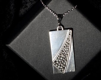 May include: A silver pendant necklace with a rectangular pendant featuring a geometric design with a textured surface.