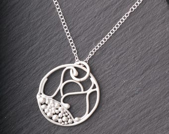 May include: A silver pendant necklace with a unique, openwork design. The pendant is circular, featuring swirling lines and a cluster of small, round beads. The necklace chain is delicate and silver, resting on a dark gray surface.