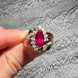 May include: A gold ring with a large, marquise-shaped ruby surrounded by smaller diamonds. The ring has a delicate, swirling design.