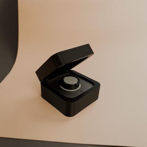 Può includere: Una scatola quadrata nera con un coperchio incernierato è aperta, rivelando un piccolo oggetto rotondo nero e argento all'interno.