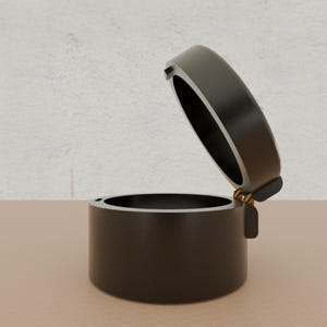 Può includere: Una scatola rotonda nera con un coperchio incernierato. Il coperchio è aperto e la scatola è vuota.