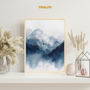 Può includere: Una stampa artistica incorniciata con un dipinto ad acquerello di una catena montuosa nei toni del blu e del bianco. L'opera d'arte è presentata in una cornice di legno chiaro. La parola "THALFO" è stampata in arancione in alto. Sono visibili vasi e piante secche.