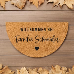 Könnte beinhalten: Eine halbkreisförmige Fußmatte mit dem Text "WILLKOMMEN BEI Familie Schneider" in schwarzer Schrift und einem schwarzen Herz. Die Matte ist hellbraun und von Herbstblättern umgeben.