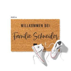 Op de afbeelding: Een bruine kokosmat met de tekst "Willkommen bei Familie Schneider" en een klein zwart hart. Twee witte hoge sneakers liggen op de mat.