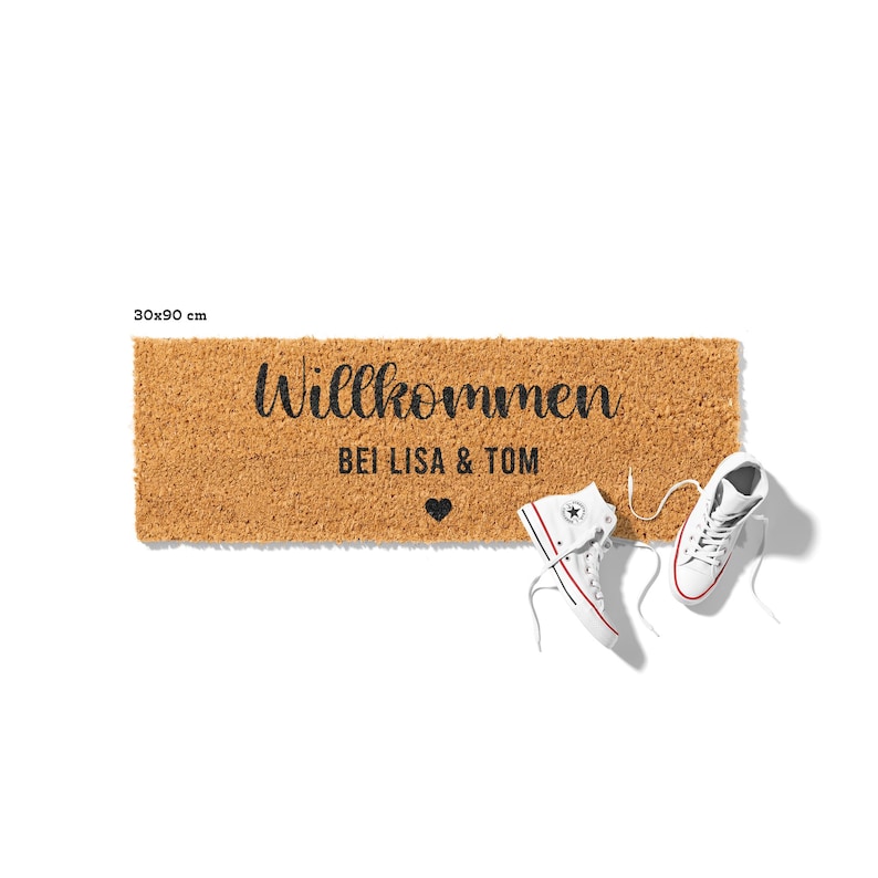 May include: A brown coir doormat with the text "Willkommen bei Lisa & Tom" and a small heart symbol. Two white sneakers are placed on the mat.