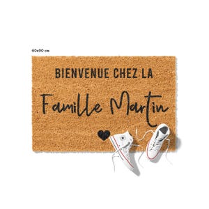May include: A rectangular coir doormat, 60 x 90 cm, with the text "BIENVENUE CHEZ LA Famille Martin" in black script. A black heart and a pair of white high-top sneakers with red and blue accents are on the mat. The mat is brown.