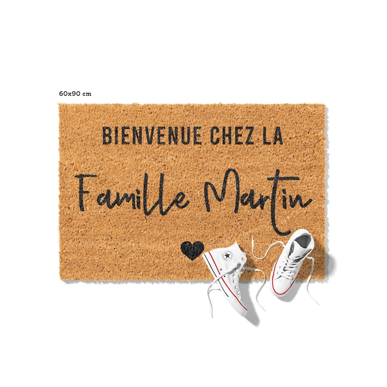 May include: A rectangular coir doormat, 60 x 90 cm, with the text "BIENVENUE CHEZ LA Famille Martin" in black script. A black heart and a pair of white high-top sneakers with red and blue accents are on the mat. The mat is brown.
