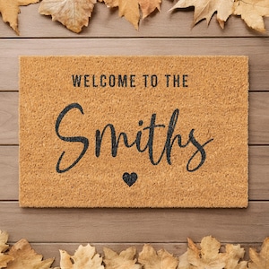 Könnte beinhalten: Rechteckige Fußmatte aus Naturkokosfaser. Der Schriftzug "WELCOME TO THE Smiths" ist in Schwarz gehalten, mit einem kleinen schwarzen Herz unter dem Namen. Braune Blätter sind um die Matte auf einem Holzgrund angeordnet.