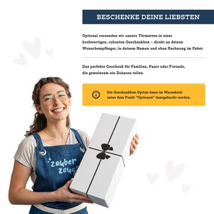 May include: A woman holding a white gift box tied with a black ribbon. The box has a black bow and is wrapped with black ribbon. The text on the box says "BESCHENKE DEINE LIEBSTEN". The woman is wearing an apron that says "zauber zeug".
