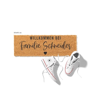 Op de afbeelding: Een bruine kokosmat met de tekst "Willkommen bei Familie Schneider" en een klein hartje in zwarte letters. De mat is 20x60 cm.