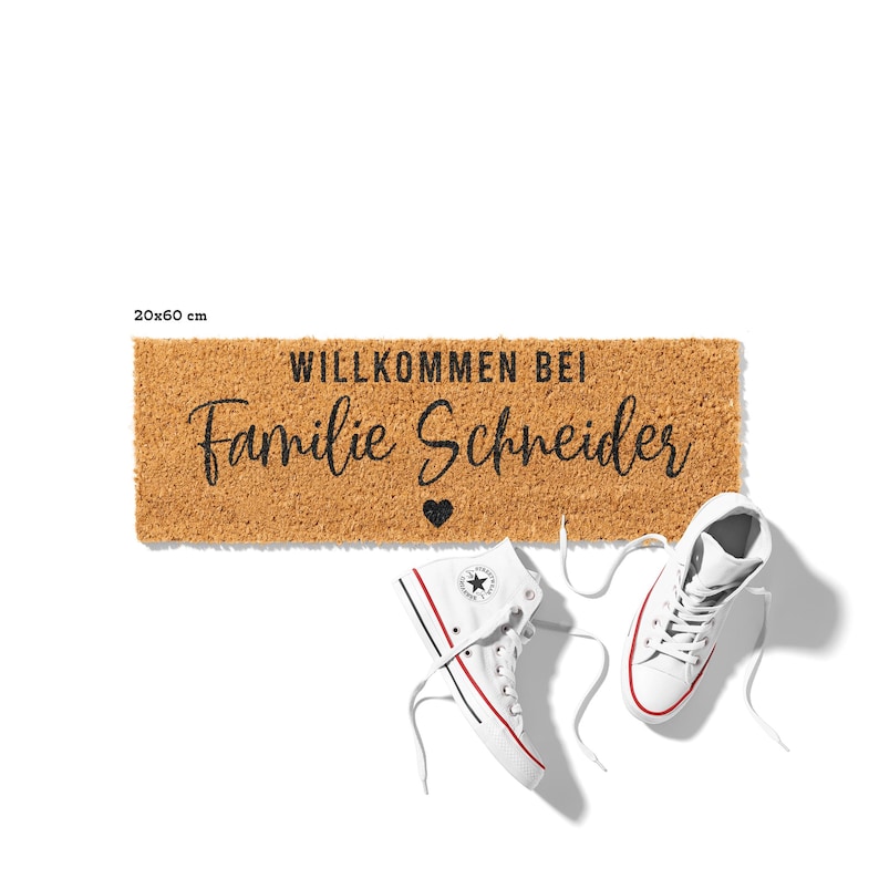 Op de afbeelding: Een bruine kokosmat met de tekst "Willkommen bei Familie Schneider" en een klein hartje in zwarte letters. De mat is 20x60 cm.