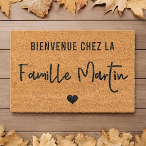 May include: A rectangular doormat with the text "BIENVENUE CHEZ LA Famille Martin" in black script, and a black heart symbol. The mat is a light brown color, with a textured surface. Autumn leaves are arranged around the mat.