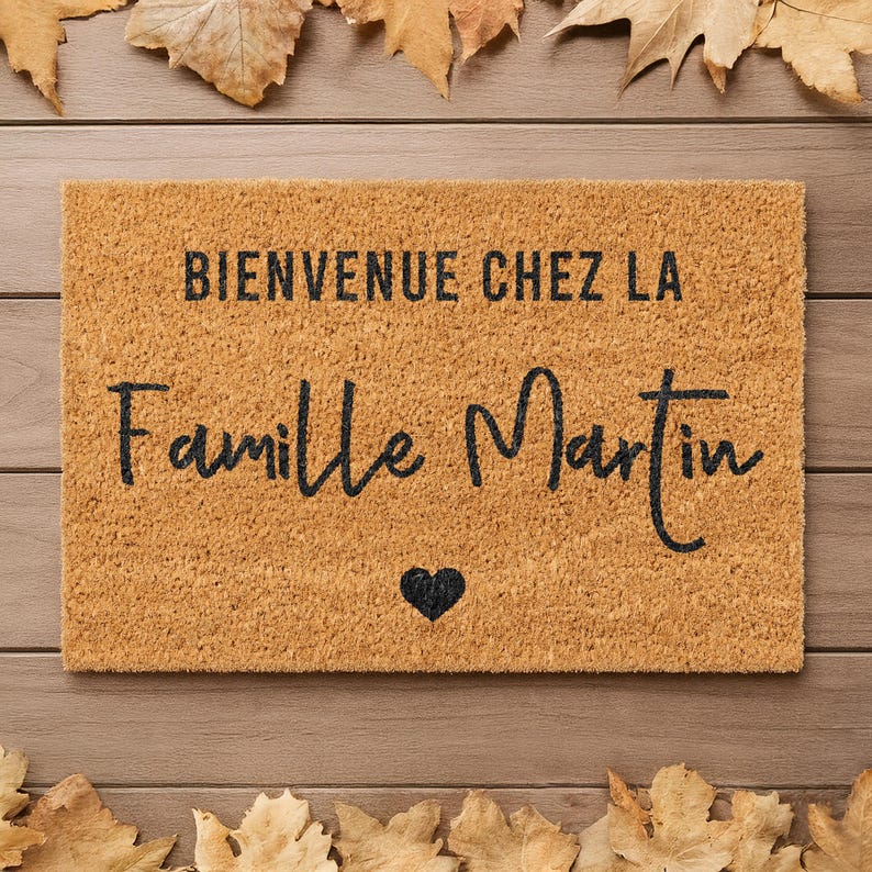 May include: A rectangular doormat with the text "BIENVENUE CHEZ LA Famille Martin" in black script, and a black heart symbol. The mat is a light brown color, with a textured surface. Autumn leaves are arranged around the mat.