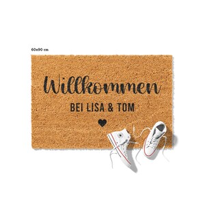 May include: A brown coir doormat with the text "Willkommen bei Lisa & Tom" and a small heart symbol. Two white sneakers are lying on the floor next to the doormat.