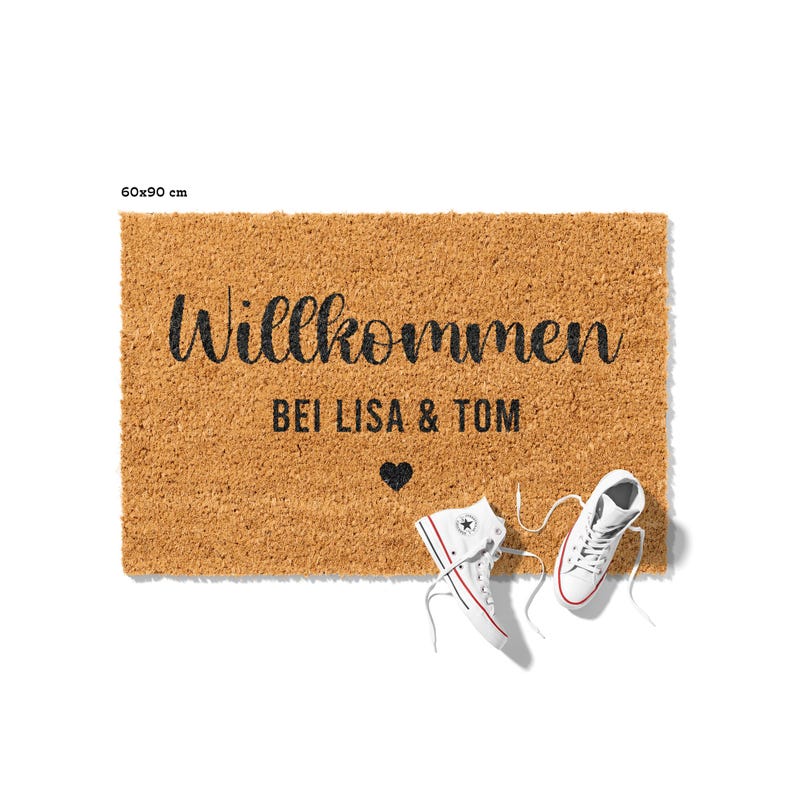 May include: A brown coir doormat with the text "Willkommen bei Lisa & Tom" and a small heart symbol. Two white sneakers are lying on the floor next to the doormat.
