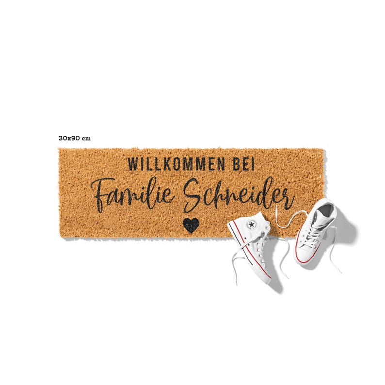 Op de afbeelding: Een bruine kokosvezel deurmat met de tekst "Willkommen bei Familie Schneider" en een klein zwart hart. Twee witte sneakers liggen op de mat.