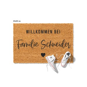 Op de afbeelding: Een bruine kokosvezel deurmat met de tekst "Willkommen bei Familie Schneider" en een klein zwart hart. Twee witte sneakers liggen op de mat.
