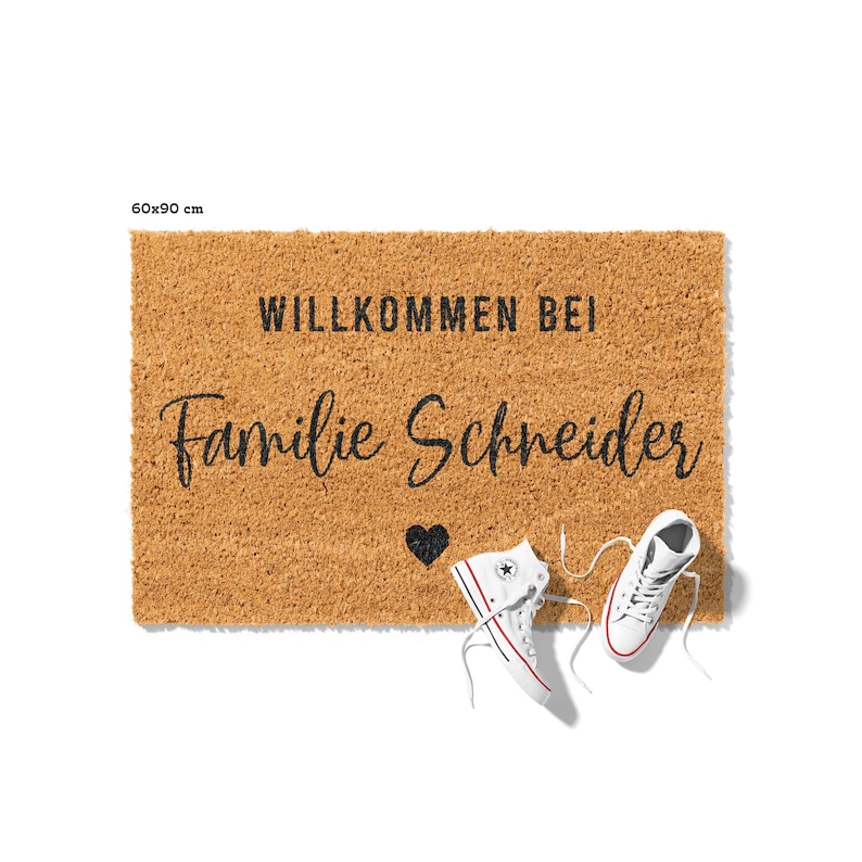 Op de afbeelding: Een bruine kokosvezel deurmat met de tekst "Willkommen bei Familie Schneider" en een klein zwart hart. Twee witte sneakers liggen op de mat.