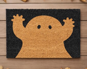 May include: A rectangular black doormat with a tan ghost design. The ghost has two black dot eyes and is raising its arms. The doormat is made of a textured material and is surrounded by autumn leaves, perfect for Halloween.