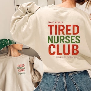 May include: A beige sweatshirt with the text "TIRED NURSES CLUB" in red and green, with "RUNNING ON CAFFEINE & CHAOS" below. A smaller version of the design is on the front. The sweatshirt is a casual garment.