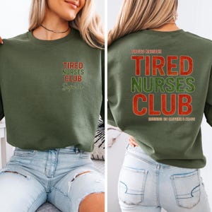 May include: An olive green sweatshirt with the text "Tired Nurses Club" in red and green. The front of the sweatshirt has the name "Sophia" below the text. The back of the sweatshirt has the text "Proud Member" above the text "Tired Nurses Club" and "Running on Caffeine & Chaos" below.