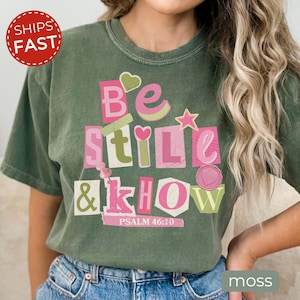Pode incluir: Uma t-shirt de manga curta verde-musgo com uma impressão gráfica com o texto "Be still & know" e "Psalm 46:10". O design utiliza um estilo de colagem com letras cor-de-rosa e verde-claro, um pequeno coração, uma estrela, um percevejo e um selo.