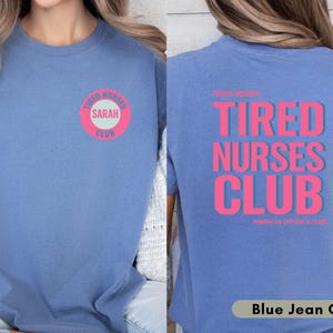 May include: Blue jean colored t-shirt with the text "TIRED NURSES CLUB" in pink on the back. The front of the shirt has a pink and white circular design that says "TIRED NURSES CLUB" and "SARAH".