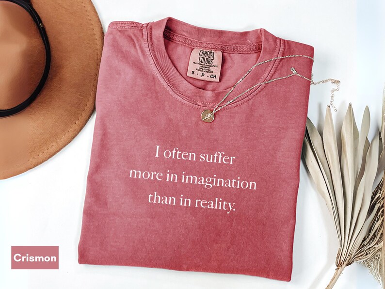 May include: Crimson-colored t-shirt with the text "I often suffer more in imagination than in reality." A gold pendant necklace and a brown hat are also in the image. The shirt is made of a soft material.