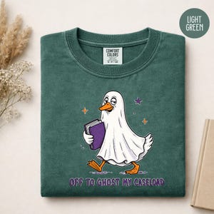 May include: A light green t-shirt featuring a graphic of a duck wearing a white ghost sheet and holding a purple book. The text below the graphic reads "OFF TO GHOST MY CASELOAD". The shirt has a Comfort Colors brand label on the inner neckline.