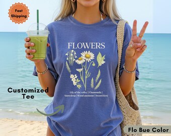 Op de afbeelding: Een blauw t-shirt met het woord "FLOWERS" in het wit, met een bloemenontwerp met madeliefjes en andere planten. De tekst "Lily of the valley | Chamomile | Snowdrop | Wood anemone | Sword fern" staat eronder. Het shirt is "Flo Bue Color".