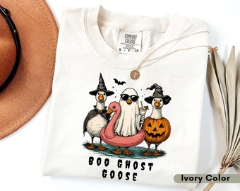 May include: Ivory-colored t-shirt featuring a Halloween-themed graphic. The design includes three cartoon geese dressed as a witch, a ghost, and a jack-o'-lantern, with the text "BOO GHOST GOOSE" below. A necklace and a hat are also visible.