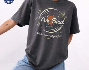 May include: A charcoal grey t-shirt featuring a graphic of a vinyl record with wings and musical notes. The text reads Free Bird, SINCE 1973, and Let the music set you free. The design is printed in cream and orange tones on the front of the shirt.