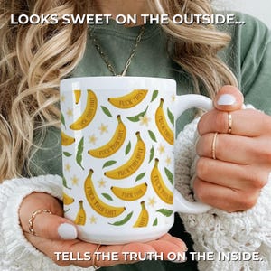 May include: A white ceramic mug featuring a repeating pattern of yellow bananas, green leaves, and small white flowers. Each banana is printed with the text "FUCK THIS SHIT". The text "LOOKS SWEET ON THE OUTSIDE..." and "TELLS THE TRUTH ON THE INSIDE." appears.
