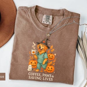 Puede incluir: Camiseta color espresso con un golden retriever con sombrero de bruja y uniforme, sosteniendo una taza de café con diseño de calabaza. La camiseta tiene el texto "COFFEE, PAWS & SAVING LIVES" y está decorada con calabazas y hojas de otoño.