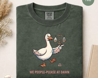 May include: A moss green crewneck t-shirt featuring a graphic of a white goose holding a balance scale. One side of the scale is labeled "MINE" and the other "THEIRS". Below the goose is the text "WE PEOPLE-PLEASE AT DAWN" and a Libra symbol.