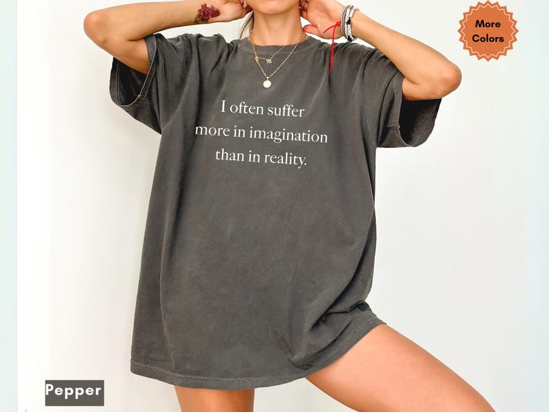 May include: A dark gray, oversized t-shirt with the text "I often suffer more in imagination than in reality" in white. The shirt has short sleeves and a relaxed fit. The word "Pepper" is at the bottom left corner, and "More Colors" is in the top right corner.