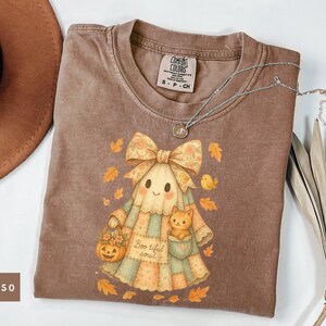 以下が含まれることがあります： エスプレッソ色のTシャツ。漫画のゴーストのデザイン、リボン、カボチャのバッグ、ポケットに猫がいます。デザインには秋の葉と「Boo-tiful soul」の文字が含まれています。シルバーのネックレスがTシャツの上にかけられています。