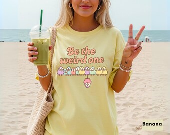 Op de afbeelding: Lichtgeel T-shirt met de tekst "Be the weird one" in een retro lettertype. Het shirt heeft een rij cartoonvogels en een roze wezen. De persoon houdt een groene drank met een rietje vast.